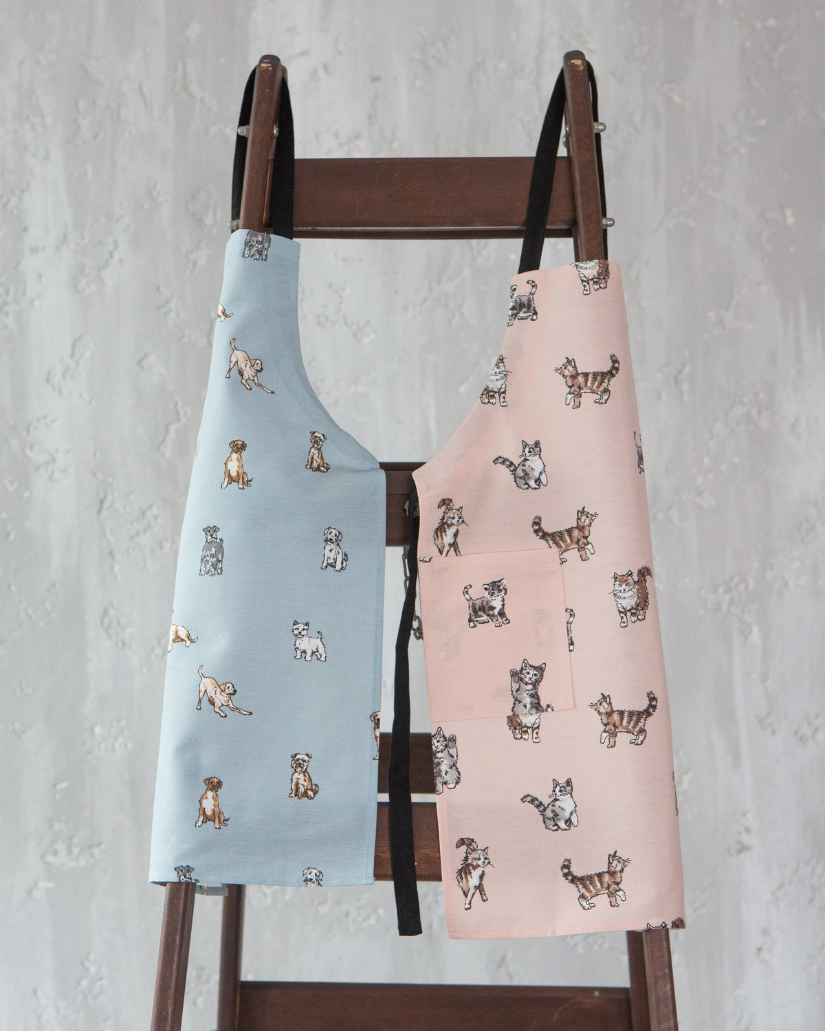 Two aprons with a cat print pattern, one light blue and one light pink, hanging side-by-side on a dark wooden ladder against a textured gray wall.